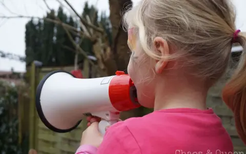 Ein Mädchen mit blonden Zöpfen hält ein Megafon und schreit hinein. Vielleicht fragt sie sich, warum sind Kinder so laut?