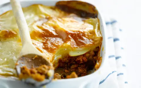 Une cuillère en bois sert une portion généreuse de moussaka végétarienne dorée et gratinée dans un plat blanc.