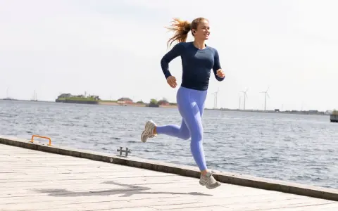 En kvinna springer glatt på en träbrygga vid havet, med vindkraftverk i bakgrunden. Hon verkar njuta av att springa 1 km varje dag.