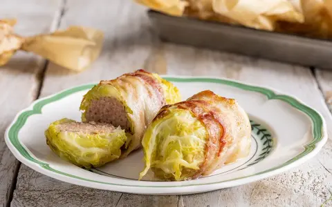 Deliziosi involtini di verza con carne, avvolti in pancetta croccante, serviti su un piatto bianco con bordo verde.