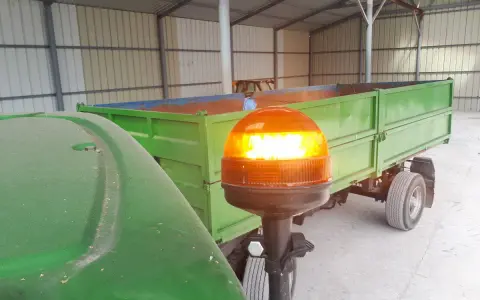 Pomarańczowa lampa ostrzegawcza na wysięgniku, przy zielonym ciągniku i przyczepie.