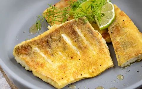Goldbraunes Zanderfilet, knusprig gebraten, serviert mit Limettenspalten und Dill auf einem grauen Teller.