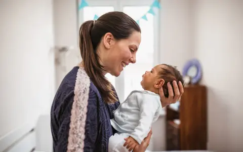Uśmiechnięta mama trzyma na rękach noworodka. Dziecko patrzy na nią z zaciekawieniem, jakby miało zaraz kichnąć.