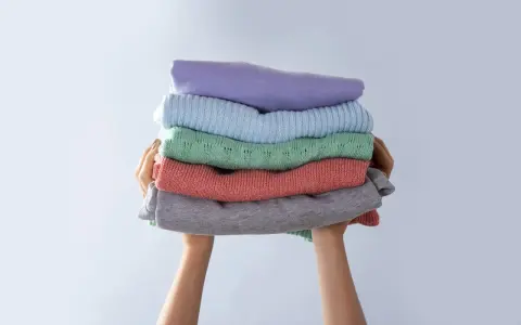 Manos sostienen una pila de ropa doblada en tonos pastel. Consejos para saber como evitar que una prenda destiña.