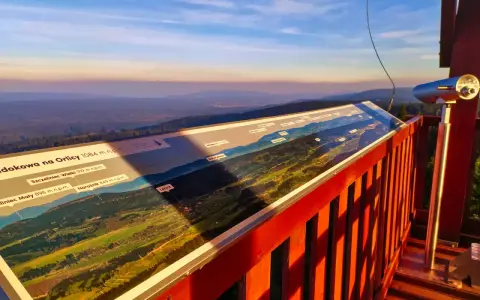 Widok z wieży widokowej na Orlicy. Na pierwszym planie panorama z opisem szczytów i dolin. W oddali łagodne wzgórza o zachodzie słońca.