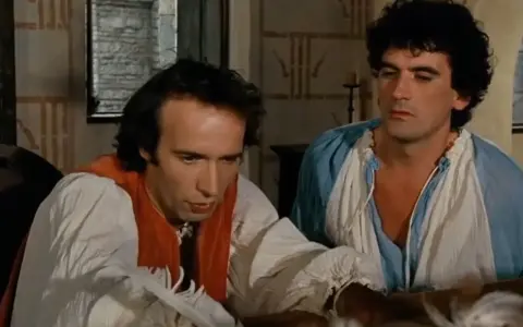 Due uomini in abiti d'epoca, uno scrive con una penna d'oca, in una scena che promette un film comico.