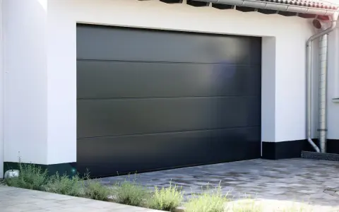 En modern, mörkgrå garageport montering med horisontella linjer. En vit fasad och grå stenläggning framför.