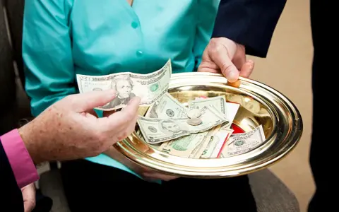 A hand places a twenty-dollar bill onto a golden tray filled with cash and coins, a gesture of offering to the poor.