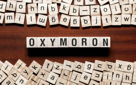 Holzbuchstaben bilden das Wort "OXYMORON". Sie sind umgeben von weiteren Buchstabenkacheln, die an Spiele wie Scrabble erinnern und Beispiele für Oxymoron-Begriffe darstellen könnten.
