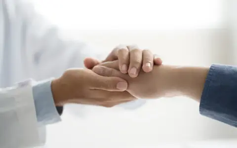 Dłoń lekarza w białym kitlu pociesza dłoń pacjenta. Symbol niepełnosprawności 02-p jest tu metaforą wsparcia.
