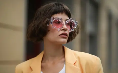 Frau mit gestuftem Bob mit Pony und auffälliger Brille mit rosa Gläsern, trägt einen hellgelben Blazer.