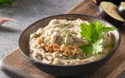 En skål med krämig baba ganoush, toppad med valnötter och persilja. Bredvid ligger skivade auberginer och ett vitlöksklyfta.