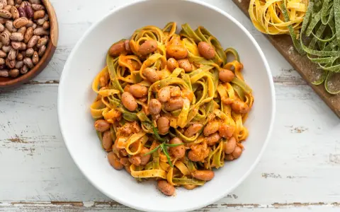 Pasta all'uovo con fagioli borlotti in una ciotola bianca, con fagioli secchi e pasta fresca colorata sullo sfondo.
