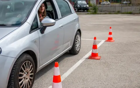 Młoda kobieta za kierownicą srebrnego samochodu ćwiczy jazdę na placu manewrowym. To może być jej droga do zdobycia prawa jazdy 17 lat.