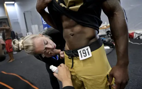 En kvinna applicerar olja på en man med en rippad kropp inför en tävling. Nummer 191 syns på hans shorts.