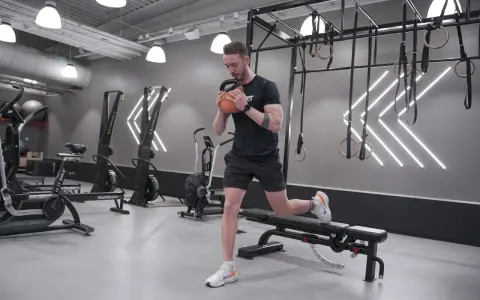 Man utför rumplyft på gym med kettlebell. Träningsredskap som roddmaskiner och ringar syns i bakgrunden.
