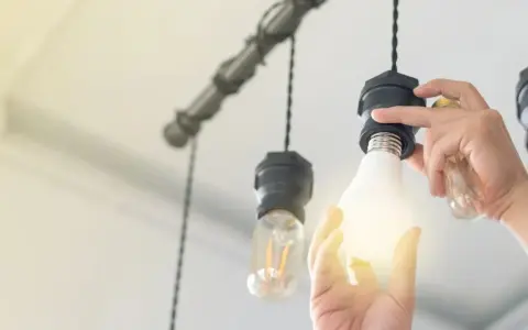 Manos cambian una bombilla LED por una incandescente en una lámpara colgante, actualizando la luz del hogar.