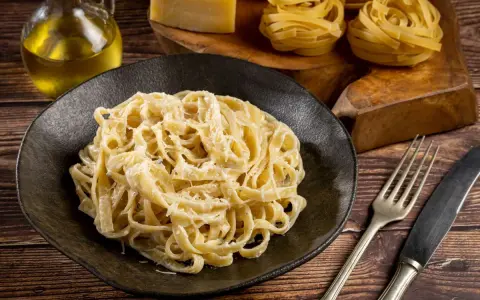 Kremowe fettuccine alfredo na czarnym talerzu, obok surowy makaron, ser i oliwa.