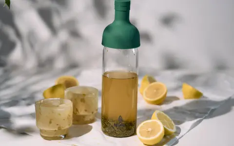 Orzeźwiający cold brew tea w butelce z zieloną zakrętką, obok szklanki z lodem i cytryny. Idealny na upalne dni.