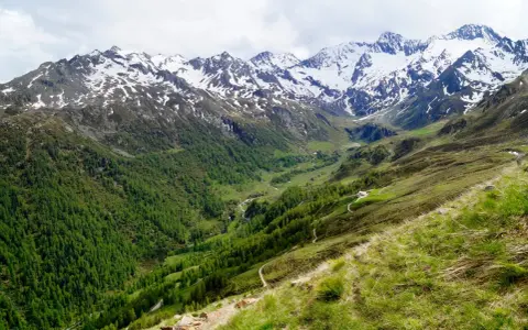 Un sentiero serpeggia su un pendio erboso verso un passo rombo in moto, con montagne innevate sullo sfondo.