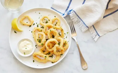 Crujientes calamares a la romana, servidos con limón y salsa tártara en un plato blanco.