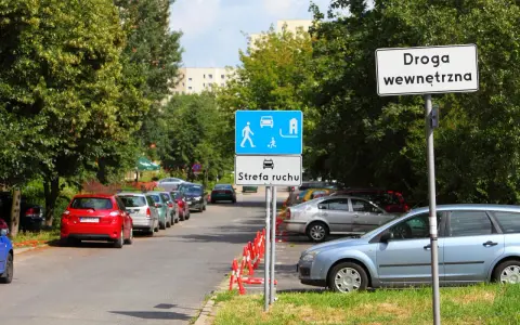 Znak "Droga wewnętrzna" obok strefy ruchu z piktogramami pieszych i samochodów. Zaparkowane samochody wzdłuż ulicy.