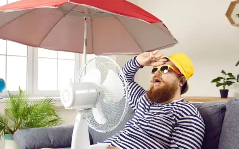 Un homme transpire sous un parasol, ventilateur et plante pour comment rafraichir sa maison. Il a chaud.