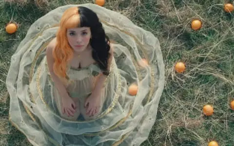Melanie Martinez, con su icónico cabello mitad naranja y mitad negro, rodeada de naranjas en un campo. ¡Atentos a los próximos eventos de Melanie Martinez!