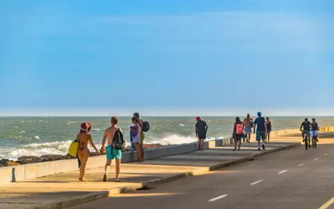 Ludzie spacerują i jeżdżą na rowerach wzdłuż promenady nad oceanem w słoneczny dzień.