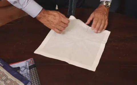 Mani esperte mostrano come piegare fazzoletto da taschino su un tavolo di legno. Altri fazzoletti colorati sono visibili a sinistra.