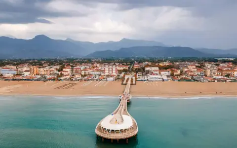 Un molo si estende su un mare azzurro, con una spiaggia dorata e una città costiera sullo sfondo. La Toscana offre un mare bello e panorami indimenticabili.