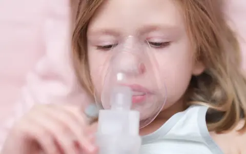 Mała dziewczynka z maską inhalacyjną na twarzy. Dziecko przechodzi inhalację na kaszel, by poczuć ulgę.