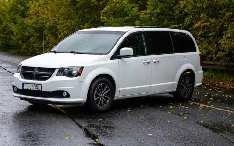Biały Dodge Grand Caravan na mokrej drodze, otoczony jesienną zielenią.