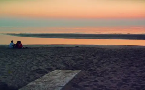 Alba a Rimini: una coppia seduta sulla sabbia guarda il mare calmo e il cielo colorato.