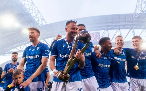 Piłkarze w niebieskich strojach świętują z pucharem i piłką na stadionie.