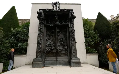 La Porte de l'Enfer de Rodin, une œuvre monumentale en bronze, se dresse au milieu d'un jardin. Deux visiteurs contemplent cette sculpture complexe.