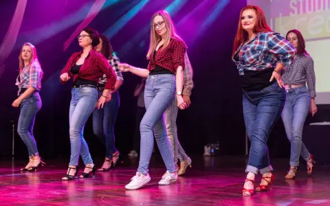 Grupa kobiet w kratę i jeansach tańczy na scenie. Występ latino solo dla początkujących.