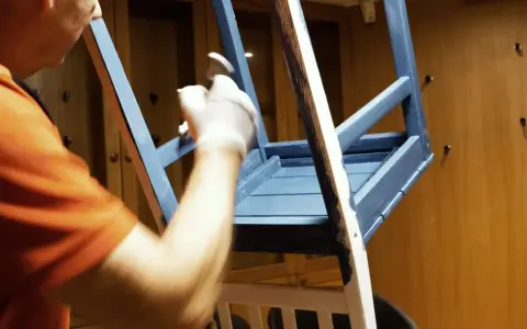 Person i orange tröja målar en blå stol med pensel.