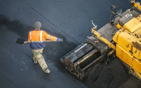 Arbeiter schaufelt heißes Bitumen auf eine Straße, während ein Asphaltfertiger das Material verteilt.