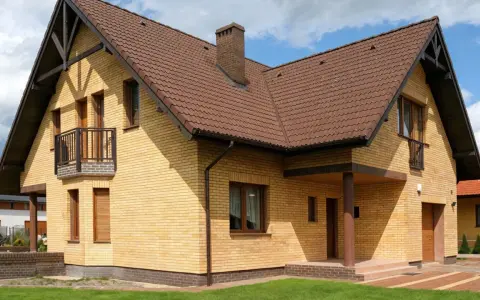 Nowy dom z żółtej cegły, z brązowym dachem i kominem. Widoczny cokół, balkon i wejście zadaszone.