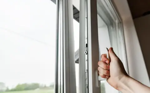 Mano girando manija de ventana blanca, asegurando la permeabilidad al aire clase 4. Ventana abierta con vista al exterior.