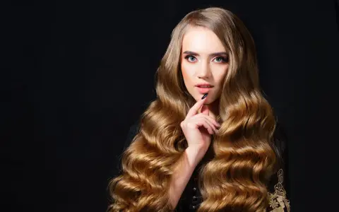 Kobieta z bujnymi, złotymi włosami ułożonymi w hollywoodzkie fale, z dłonią przy ustach, na czarnym tle.