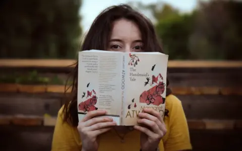 Młoda kobieta w żółtej bluzce czyta "Opowieść podręcznej" Margaret Atwood. Jej oczy błyszczą, gdy zagłębia się w świat literatury, co jest idealnym przykładem pasji do filologii.