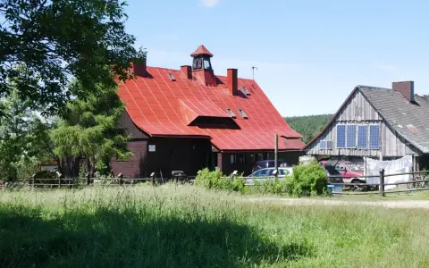 Chatka Górzystów z czerwonym dachem i drewnianymi budynkami. W tle las, a przed budynkami zielona łąka.