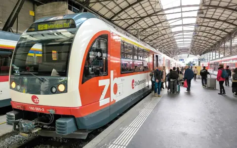 A train labeled "PE Interlaken, Ost" waits at the station, ready for its journey on the Lucerne to Interlaken Golden Pass line. Passengers gather on the platform.