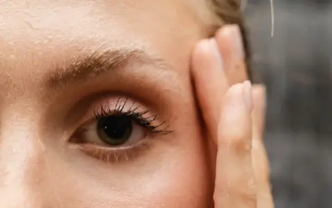 Un œil attentif, cils longs, et le début du repousse sourcils après chimio. Une main effleure le visage.