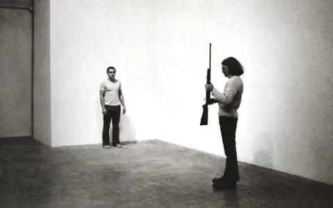 Chris Burden, dans une performance tendue, tient un fusil. Un homme se tient à distance, le regard fixé.