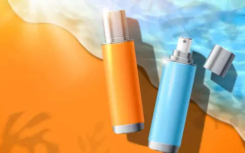Due flaconi spray, uno arancione e uno azzurro, sulla sabbia dorata con onde marine. Protezione solare con butyl methoxydibenzoylmethane.