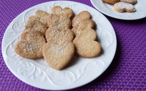 Stos ciasteczek cytrynowych w kształcie serc, posypanych cukrem pudrem, na białym talerzu.