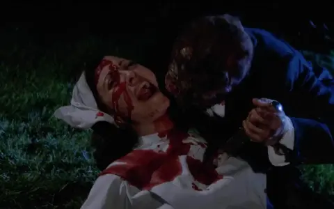 Una vittima urlante, coperta di sangue, viene attaccata da una figura grottesca con un coltello. Un'immagine raccapricciante che evoca un **horror cult**.